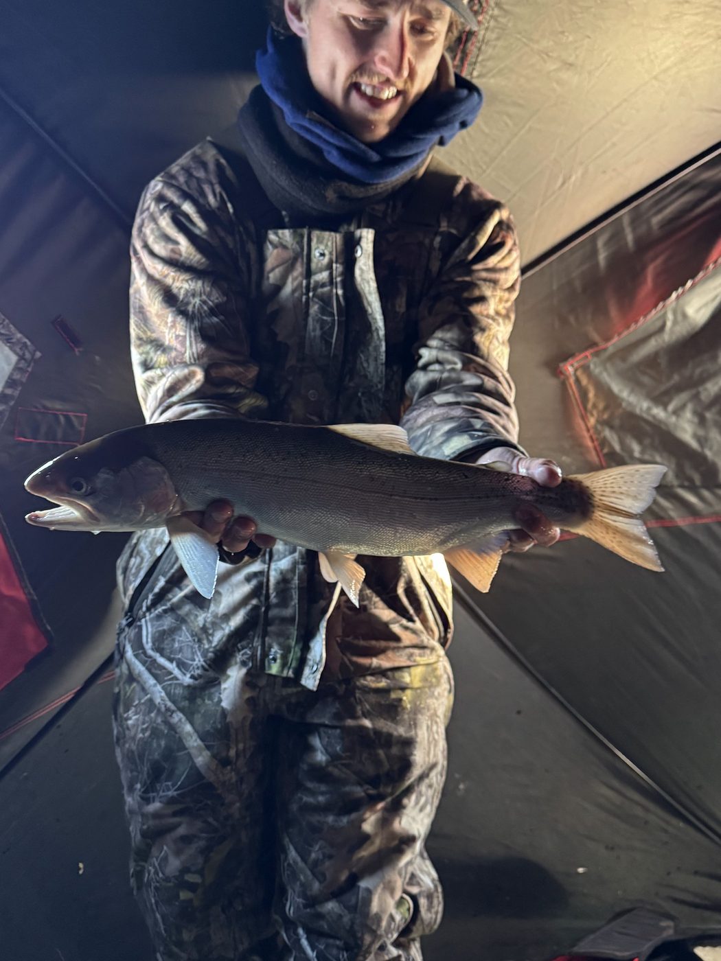 Angler in camouflage holding a large rainbow trout inside an ice shelter