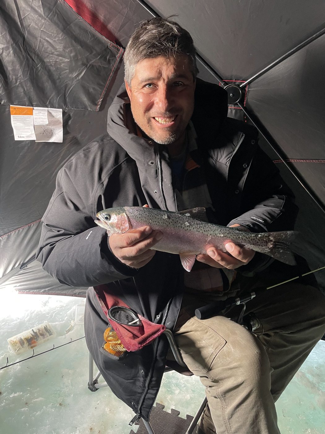 Angler in a dark jacket holding a rainbow trout inside an ice shelter