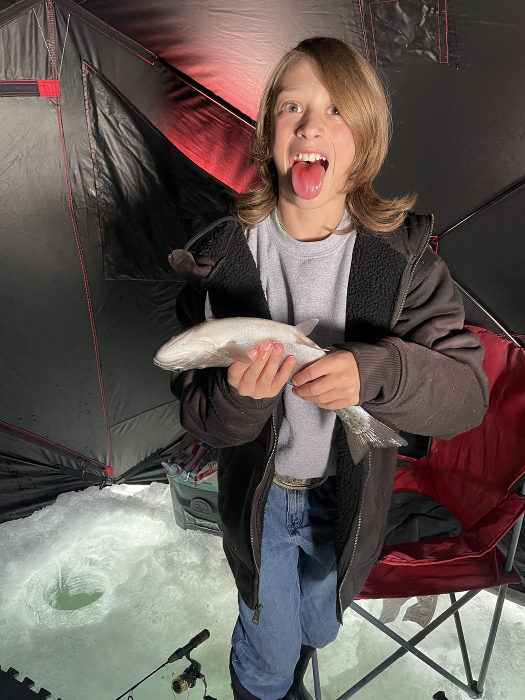 Young angler making a funny face while holding a trout inside an ice shelter