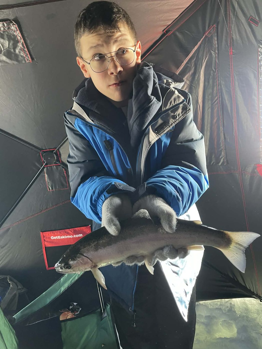 Young angler in a blue jacket and gloves holding a trout inside an ice shelter