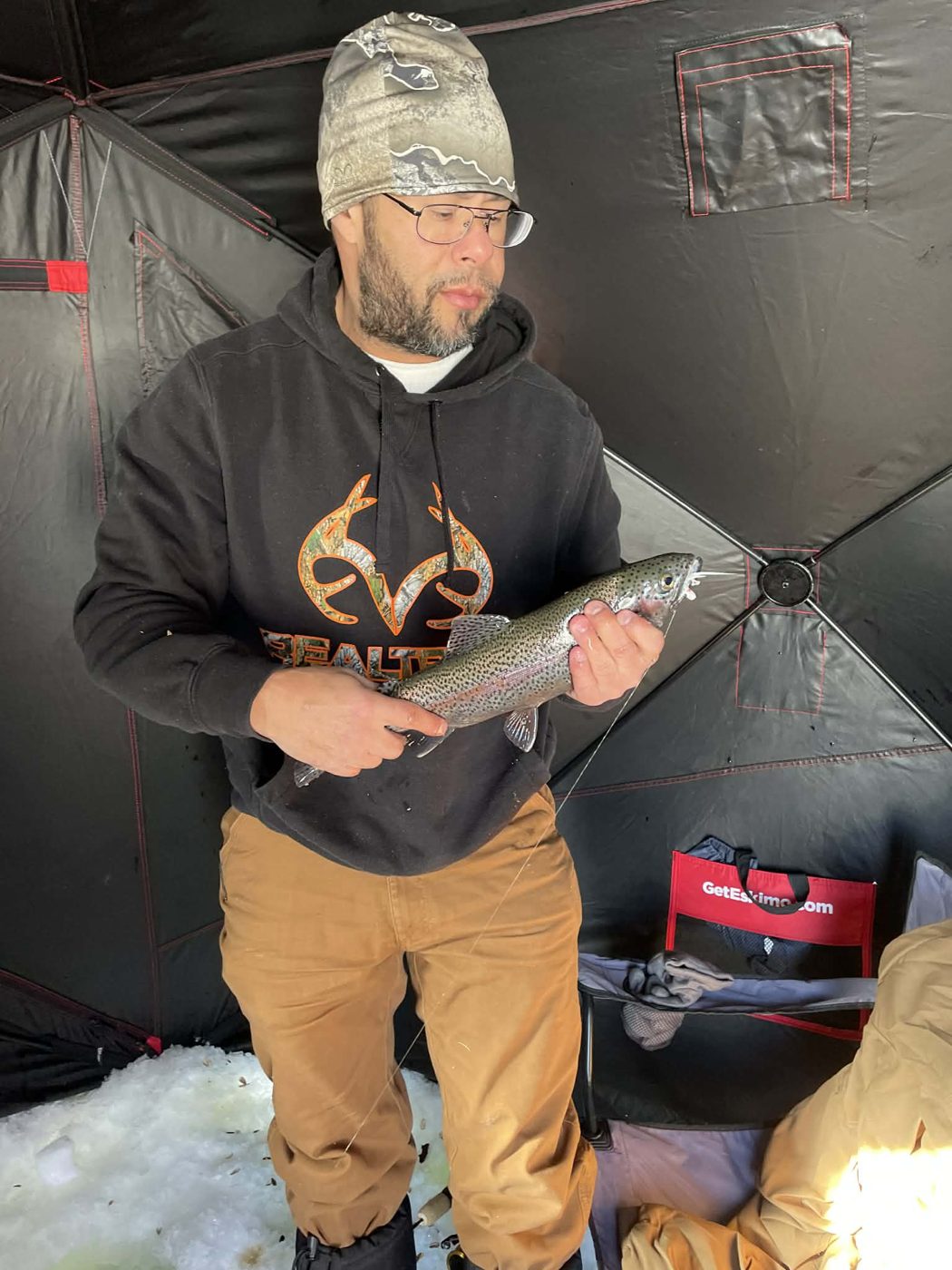 Angler in a camouflage beanie and black hoodie holding a rainbow trout inside an ice shelter