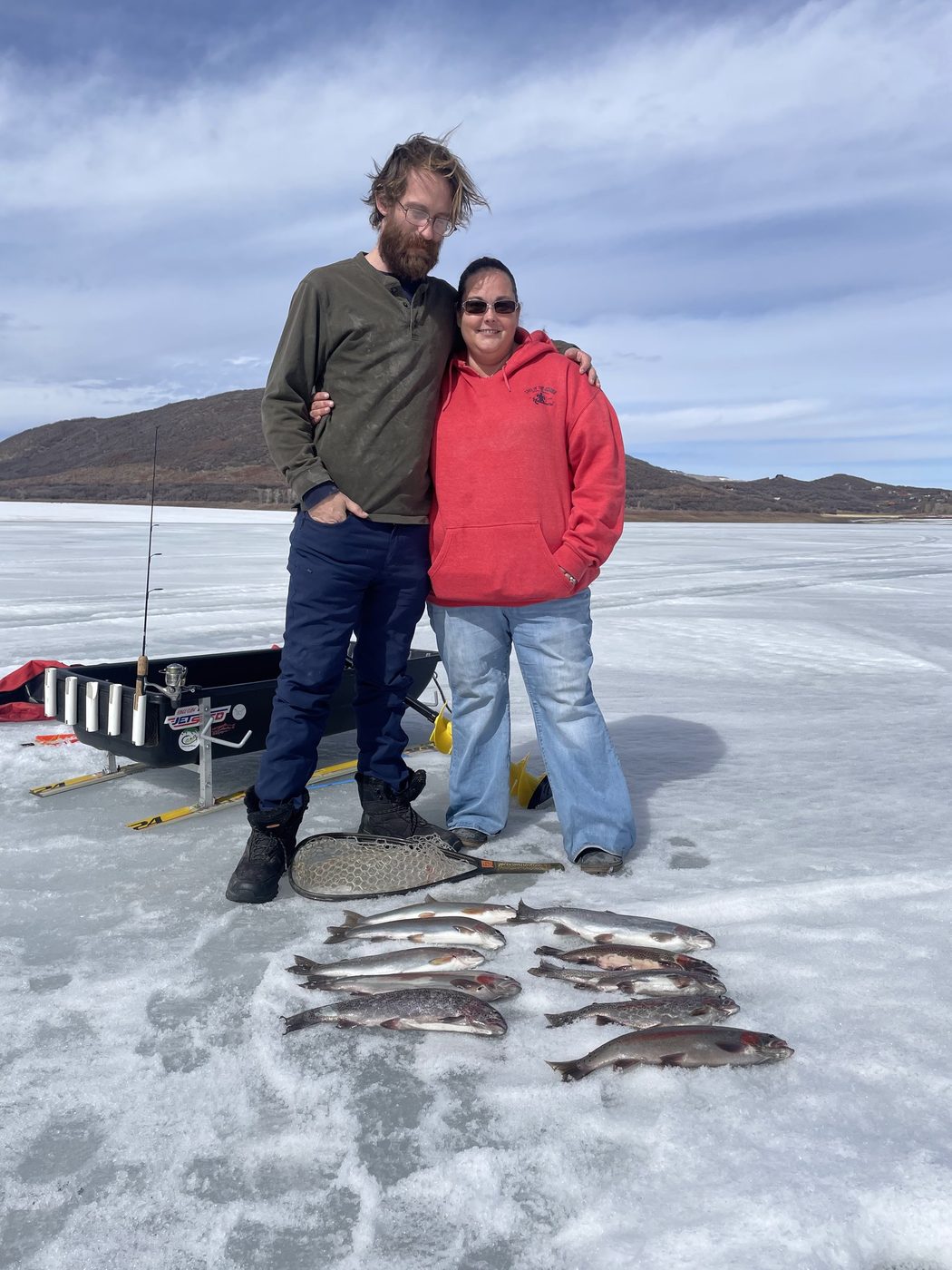 Two anglers standing on a frozen lake with a row of freshly caught trout laid out on the ice