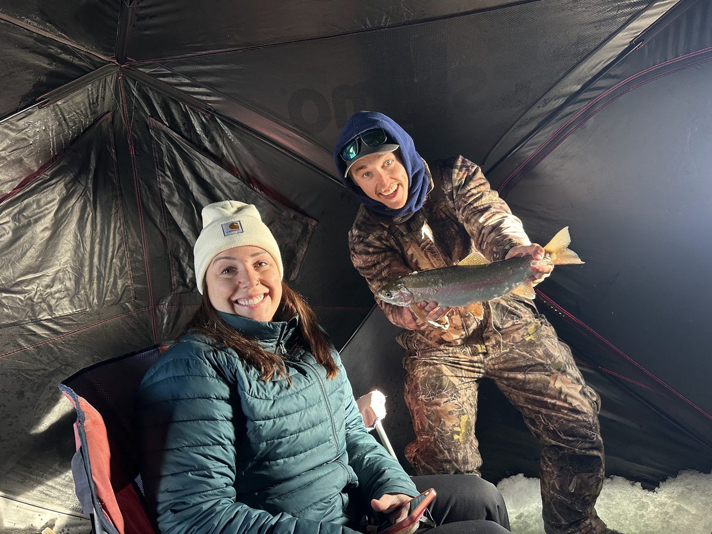 Two guests smiling inside an ice fishing shelter, one holding a rainbow trout