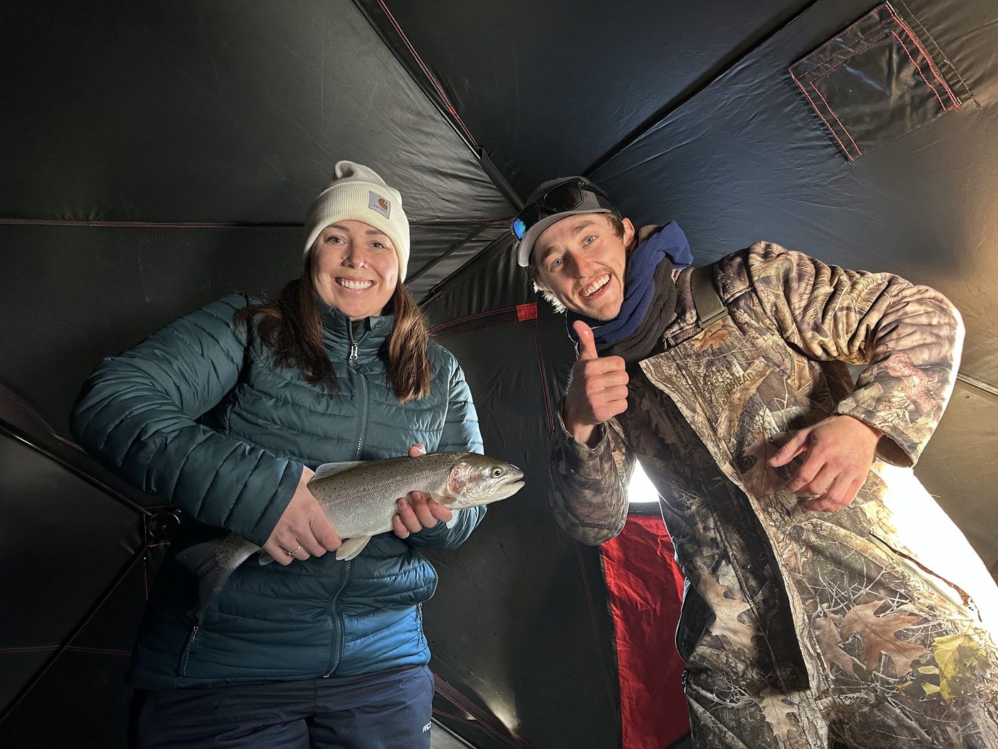 Two guests posing with a trout inside an ice fishing shelter