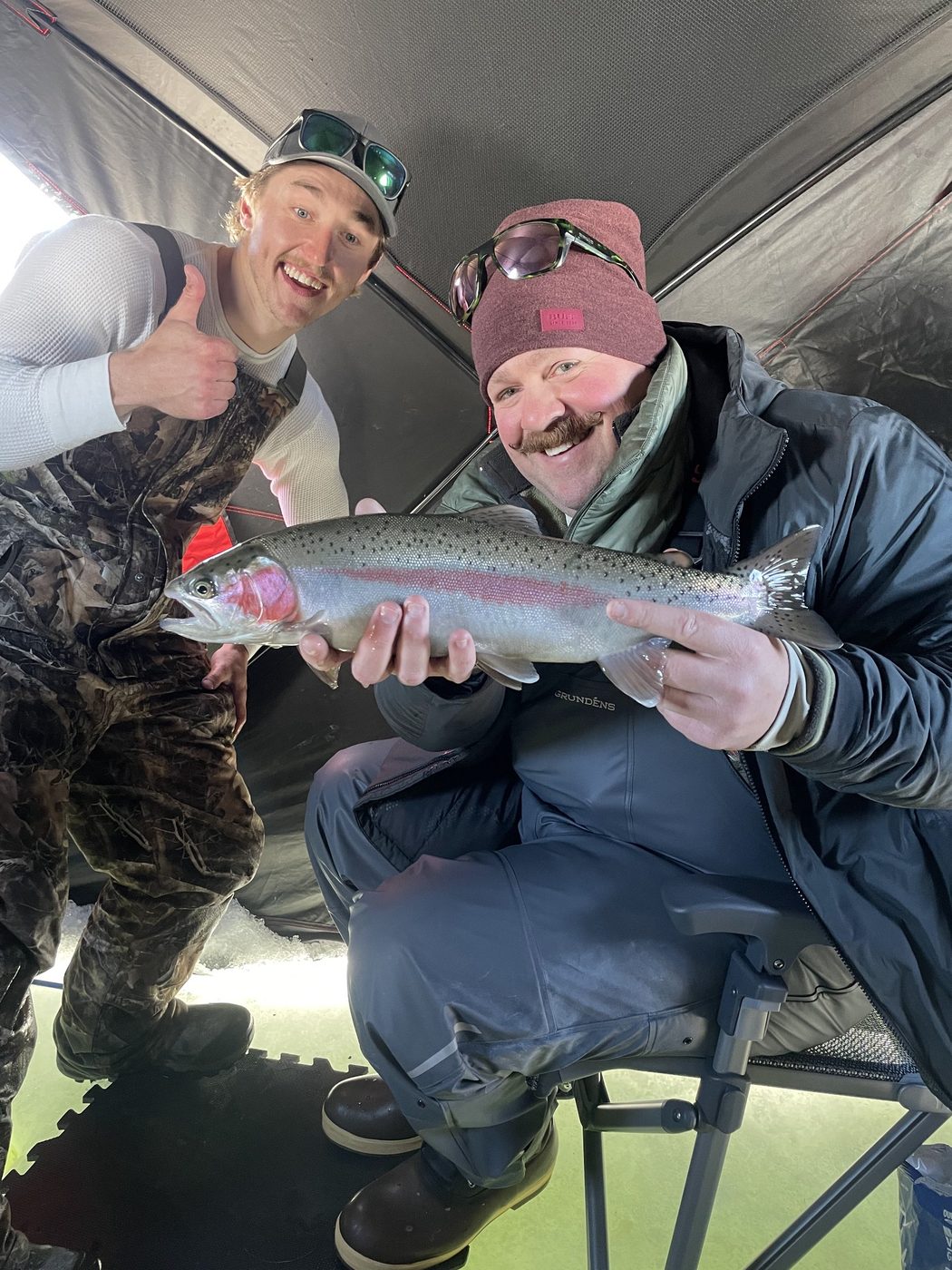 Two anglers inside an ice fishing shelter holding a rainbow trout