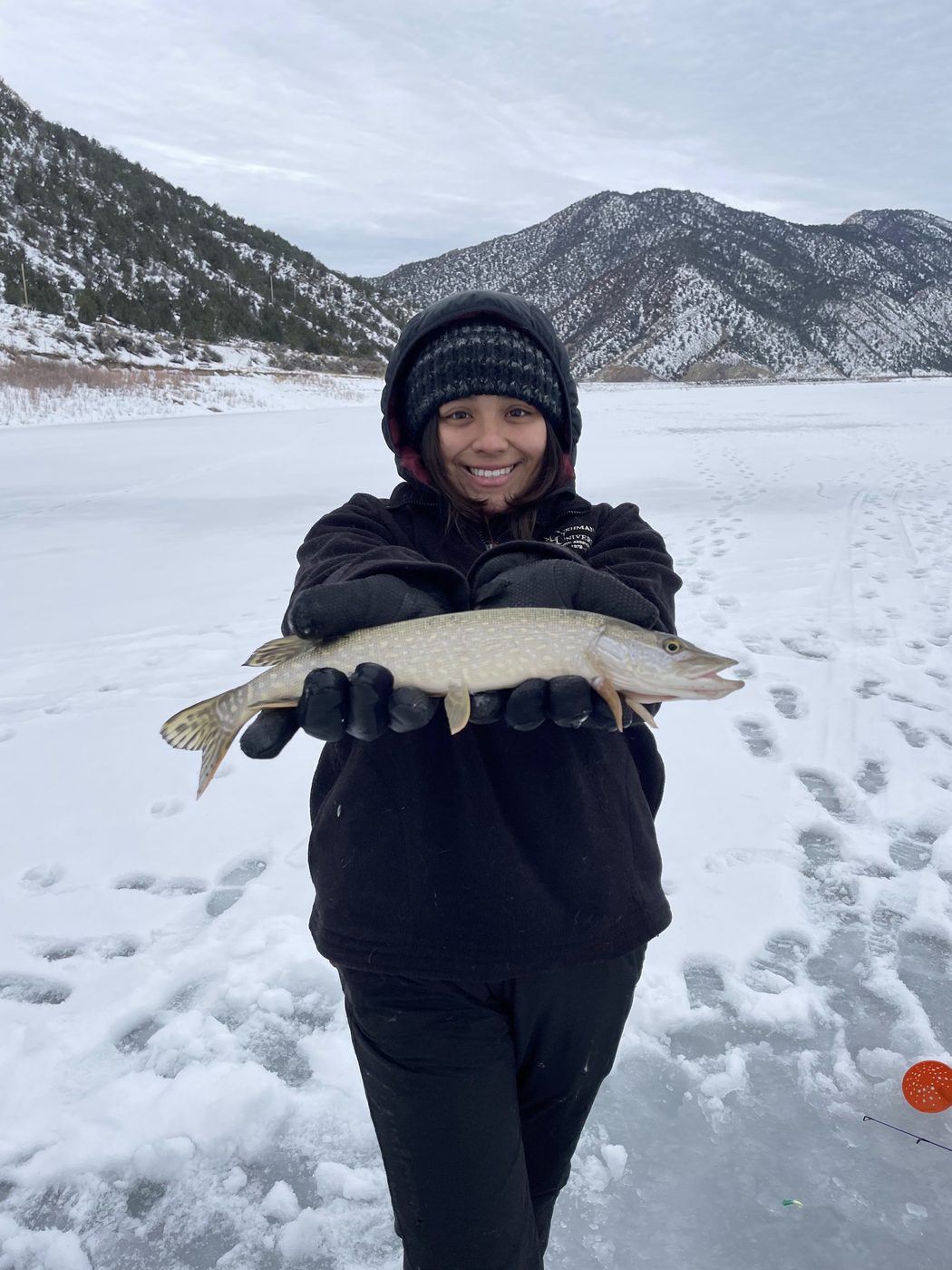 Smiling angler holding a northern pike on a frozen lake with snowy mountains behind